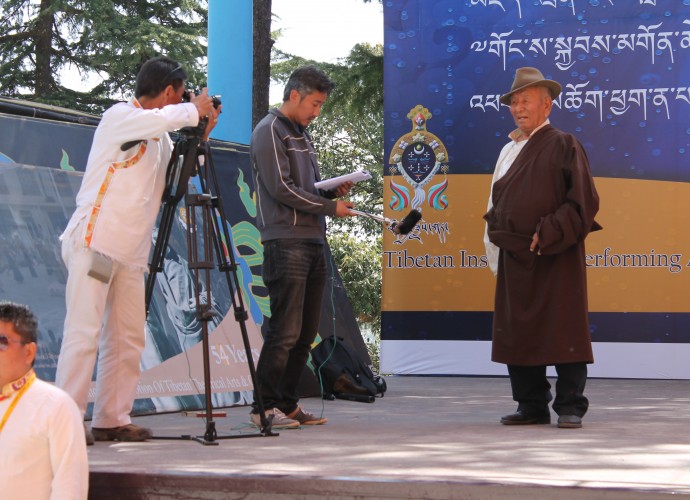<p>Pema Tashi (cameraman), Sonam Wangdue (interviewer) and Kundeling Thupten (performer)<br />
McLeod Ganj, Dharamshala, India <br />
25 March 2015 <br />
Photographer, Emma Martin</p>
<p>As part of NML’s collecting project <em>Tibetan Realities: Collecting Tibet and its Diasporas,</em> the author attended the 20th Shoton (Tib. zho ston) festival held at McLeod Ganj, Dharamshala in Himachal Pradesh in March/April 2015. Over the course of two weeks 12 troupes from across South Asia came to perform traditional and newly commissioned operas at the Tibetan Institute of Performing Arts (TIPA). I worked with Tibetan cameramen and producers Pema Tashi and Sonam Wangdue to record this major event for inclusion in the NML exhibition, <em>Tibet: Dreams and Realities</em>. Here, Pema Tashi and Sonam Wangdue are conducting an interview with acclaimed <em>ache lhamo</em> performer, Kundeling Thupten. 82-year-old Kundeling Thupten formed an <em>ache lhamo</em> troupe in 1959 after escaping into exile. He lives in Bylakuppe, South India and still performs for the Bylakuppe Lhamo Association.</p>