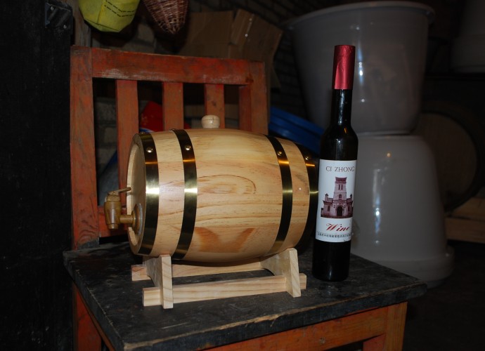 <b><p>Wu Gongdi’s and his son Hongxing’s wine in a barrel and a Cizhong labeled bottle.</b></p>
Most households in Cizhong produce relatively small amounts of wine compared to Wu Gongdi and only sell to visitors and also sell some grapes to the local Shangri-La Wine Company. The Shangri-La Wine Company is a recent development throughout the lowland river valleys of Deqin County where Cizhong is located. Developed in the early 2000’s by the local government as a poverty alleviation scheme, the company and the county government have promoted the conversion of traditional agricultural fields of barley and wheat throughout Deqin into monocroped vineyards. In most villages the grapes produced in these vineyards are sold directly to the company for winemaking rather than being used for household winemaking as they are in Cizhong. While the growing of grapes has overall led to higher annual incomes for households throughout the region, there have been drawbacks in terms of sustainability and food security. Having abandoned the production of grains for subsistence consumption, households are reliant on the sale of grapes to purchase food, and the Shangri-La Wine company does not have guaranteed contracts with villagers from one year to the next, often showing up late to purchase grapes after they have ripened and begun to dry out. In addition, the grapes introduced to Cizhong more recently and to other villages by the government and the company are of a different Cabernet variety, which requires heavy use of chemical pesticides and fertilizers. For more detailed information and these agricultural transformations in Deqin County associated with viticulture, see (Galipeau 2015).
