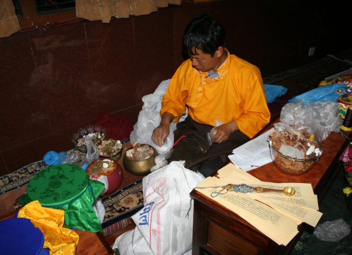 Amchi Nyima is mixing the raw ingredients and filling the cardinal medicinal vessels to be placed upon the mendrup mandala during the ritual. He closely follows the photocopied manuscript of the mendrup medicinal formula which lists more than one hundred ingredients and is placed on the table in front of him. Amchi Nyima is including all of those herbal ingredients that he can comprehend from the formula, as well as a few minerals and one animal substance (red lac). Nevertheless, he skips the part of the recipe listing animal and human body parts. The colorful ritual ‘spell cord’ (byang thag, gzungs thag) is wrapped around a ceremonial brass ladle. The spell cord has five strings in the five colors of the cardinal directions. The cord connects the abbot presiding over the ritual with each point on the mandala and its medicinal vessel (see below). The ladle will be used for liquid offerings to the deities invoked. 
