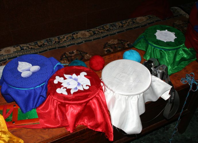 The medicinal vessels of the five cardinal points are covered with cloth lids of their respective signs and colors: the white cloth with a swastika will occupy the Centre, the green with a dharma wheel the North, the red embellished with a lotus flower will represent the West, and the blue with a flaming jewel the South. The yellow cloth covered bowl representing the East is missing in the photograph.