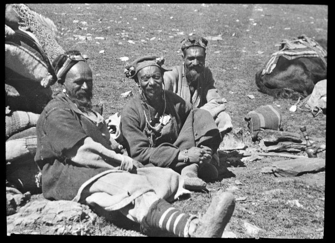 From their distinctive headgear, we can see that these men belong to one of the Brokpa (’Brog pa, ‘Dard’) villages of Dah, Hanu, or Garkhon, which today lie close to the Line of Control between Indian- and Pakistani-administered territory. The Brokpa speak a distinct language that belongs to the Indo-European group of languages rather than a Tibetan-related dialect. These men are Buddhists, as we can see from the amulet worn by the man in the middle, but some neighbouring Brokpa communities are Muslim.
It seems as though they are resting in the course of a journey. To the right we can see a ‘hookah’ pipe for smoking tobacco. They may have been carrying grain submitted to a nearby regional centre as a form of taxation.
