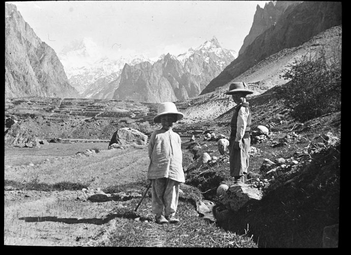 Our final image shows the Reads’ two children, John and Marguerite. Each child is wearing a solar topi, a form of protection that was then considered essential, especially in the more intense sunlight that one encounters at high altitudes.
The scene is near the Reads’ home in Khapalu. In the background we can see the Hushe valley together with the Masherbrum mountain peak. The picture must have been taken in the early spring because we can see the first blades of wheat sprouting up in the field next to the two children.
The small ridges in the field are designed to assist irrigation. In both Baltistan and Ladakh the distribution of water from mountain streams is carefully regulated by village leaders.
The two children grew up speaking Balti and Urdu and, many decades later, John Read still remembers many of the words in these languages that he learnt as a child.