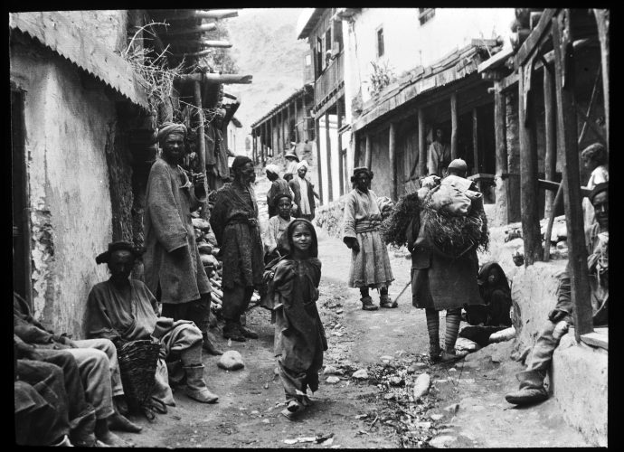 This scene is from Kargil, Ladakh’s second town, which is approximately the same distance—eight days’ march in earlier times—from Leh, Srinagar and Skardu (Baltistan). The majority of its inhabitants are Shia Muslims.
To judge by their clothing, the people in this image represent a variety of different groups. The man facing the camera in the middle distance may be a Buddhist. The figures behind him are wearing white turbans and may be traders from the Indian plains.
Note the girl’s bare feet. She’s not the only one to have no shoes. One of the men sitting on the left—we can only see his legs—is in a similar condition.