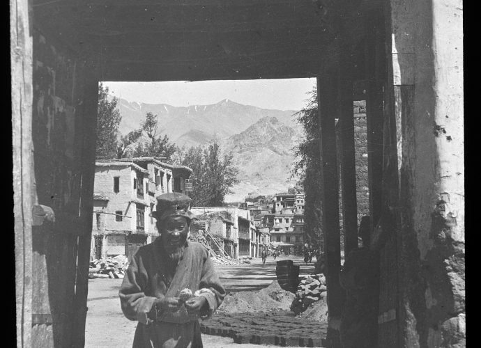 This is the Balkhang gate at the entrance to Leh bazaar, which was demolished in the 1970s. The Jama Masjid, the main mosque of Leh, which dates back to the mid-17th century, is visible at the far end of the bazaar. The old man appears to be spinning wool as he walks.
In the middle distance, we can see sun-dried bricks laid out in preparation for building work. On the left there is a pile of stones, presumably set aside for the same purpose. The British army officer Rupert Wilmot (Wilmot et al. 2014: 40) took a similar picture in 1931.
