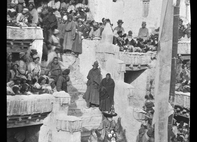 This scene is from Spituk (dPe thub) monastery, five miles from Leh, and shows part of the annual Gustor (dgu gtor) festival which takes place in the 11th month of the Tibetan year (Marko 1994). Over two days the Spituk monks perform cham (’cham) religious dances. A monk dressed in elaborate ceremonial robes is about to descend from the main temple into the courtyard for the Black Hat Dance (zhwa nag rnga’cham). On all sides crowds of villagers wait expectantly. To the right one can see the tall mast that stands in the center of all monastic courtyards, with a long prayer flag hanging from it.